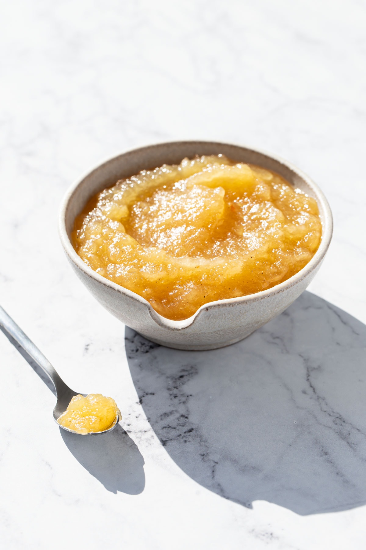 Spouted ceramic bowl filled with a swirl of homemade applesauce, with a spoon of applesauce sitting on the side.