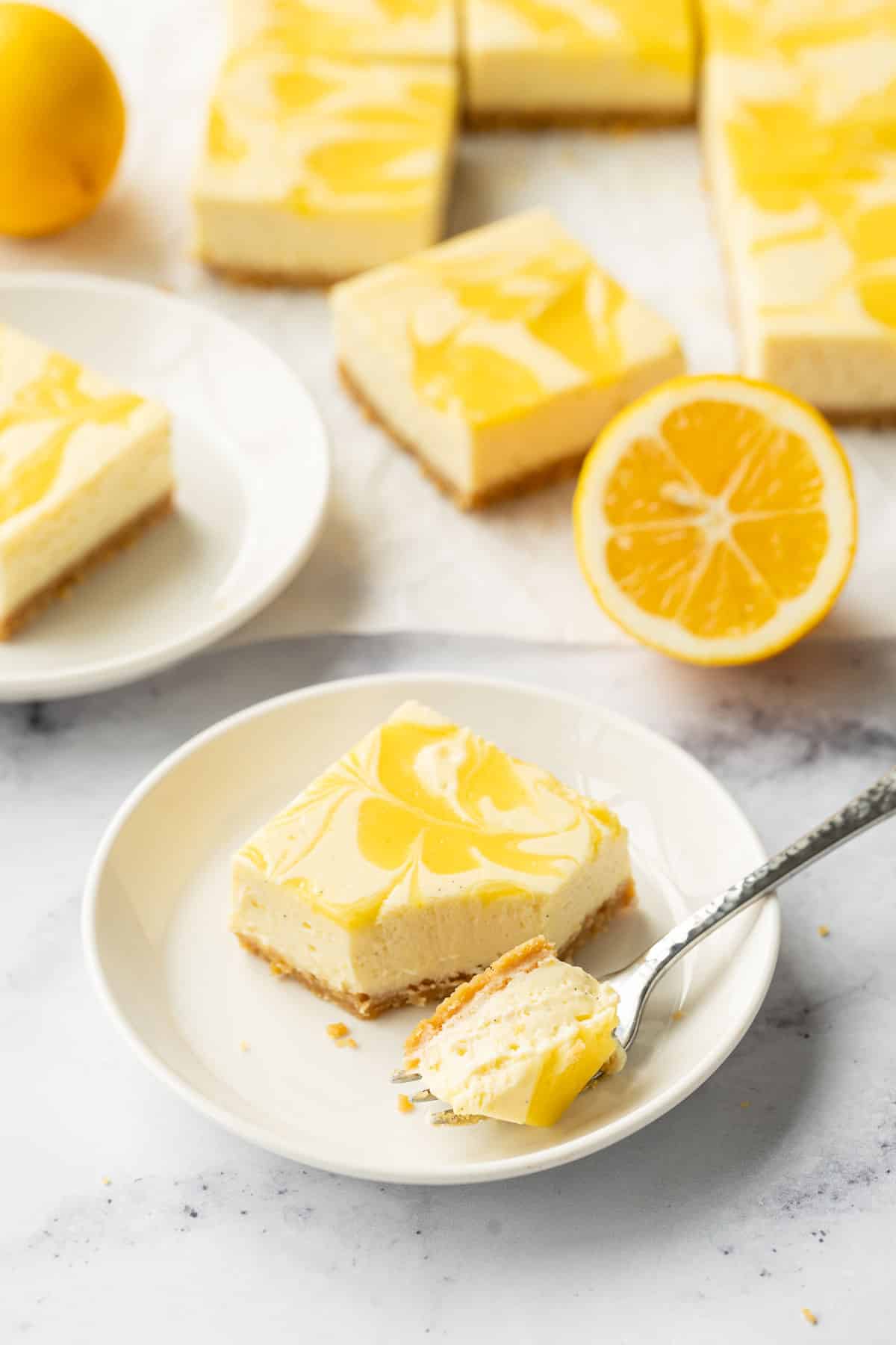 Square of Meyer Lemon Cheesecake on a small white plate with a forkful cut out to show the texture, more cut bars and a few lemons in the background.