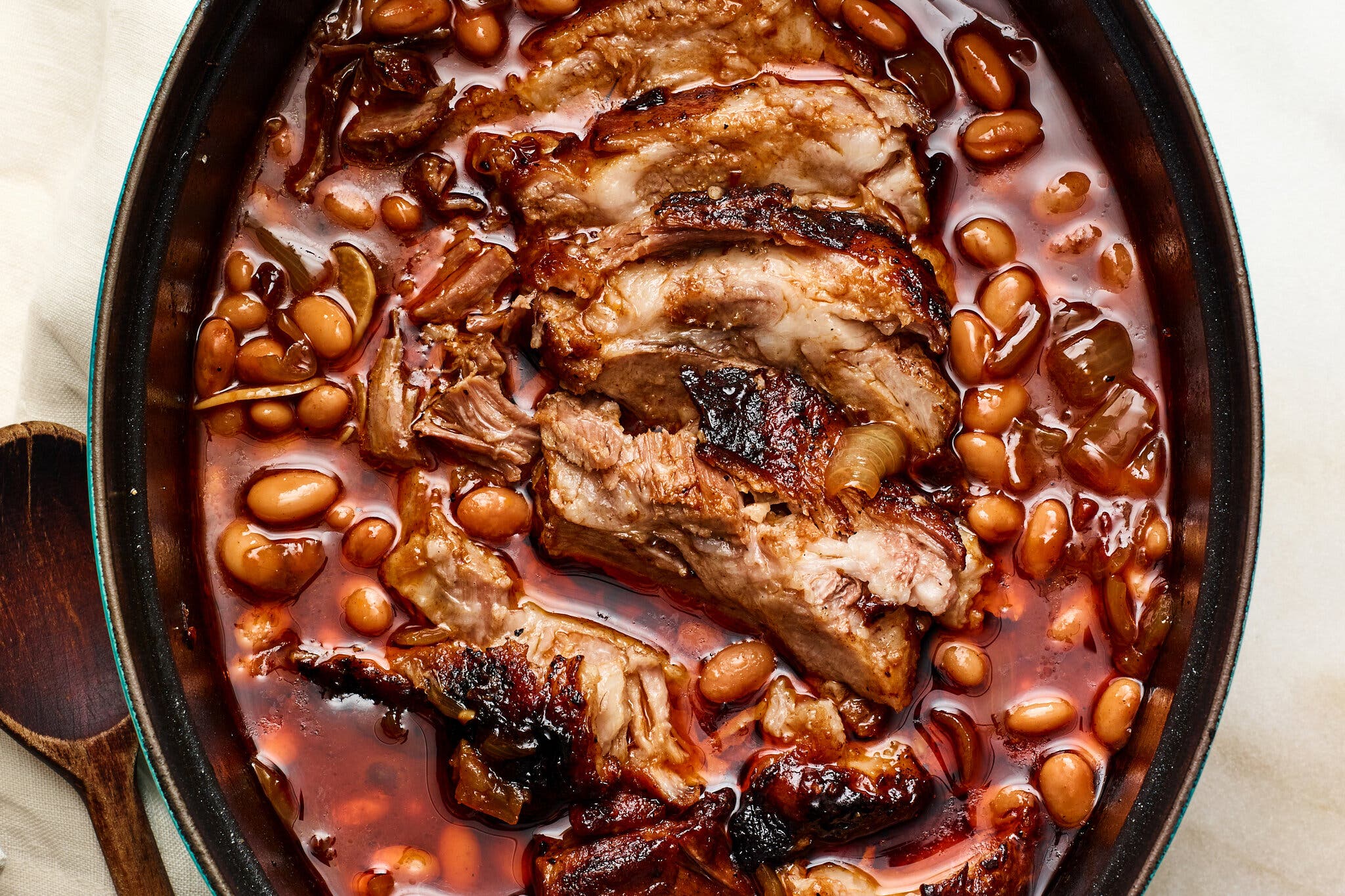 An overhead image of an black oval casserole dish filled with kidney beans and sliced, burnished pork roast in a brick red broth.