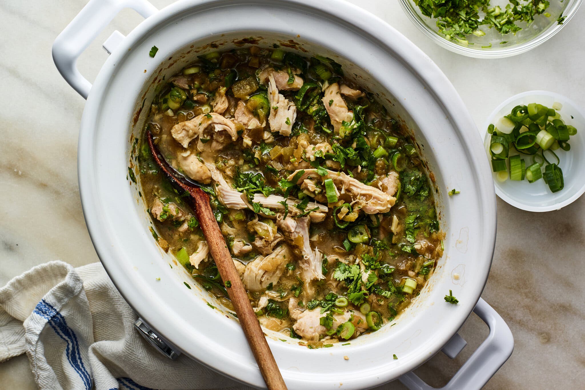 A white slow cooker holds a brothy stew strewn with green herbs and chunks of chicken. A small bowl with scallions and a slightly larger bowl with herbs sit off to the side, and a gently rumpled tea towel to the other side.