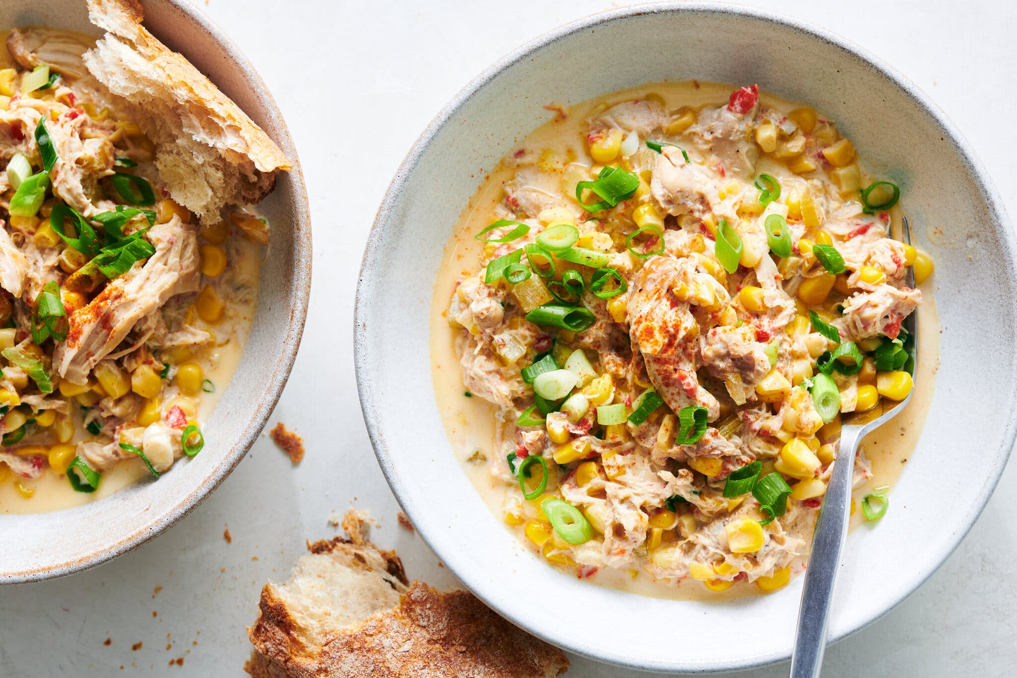 An overhead shot of two bowls of creamy chicken and corn stew, garnished with chopped green onions, with pieces of bread on the side.