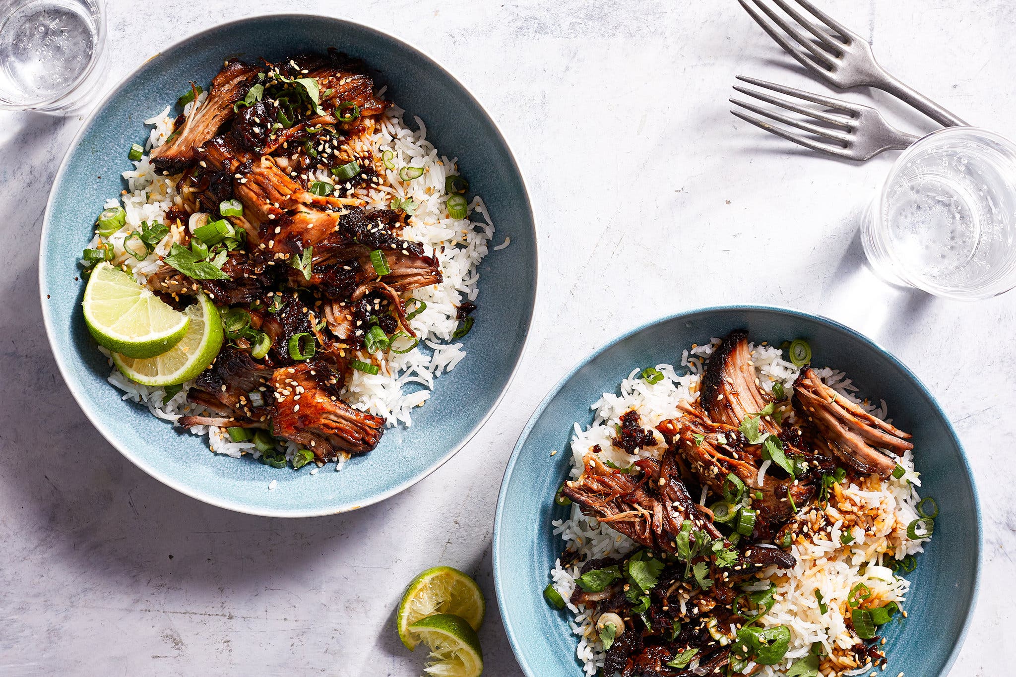 An overhead image of two blue bowls filled with rice and topped with shredded glazed pork, chopped scallions and sesame seeds. Lime wedges sit on the side.