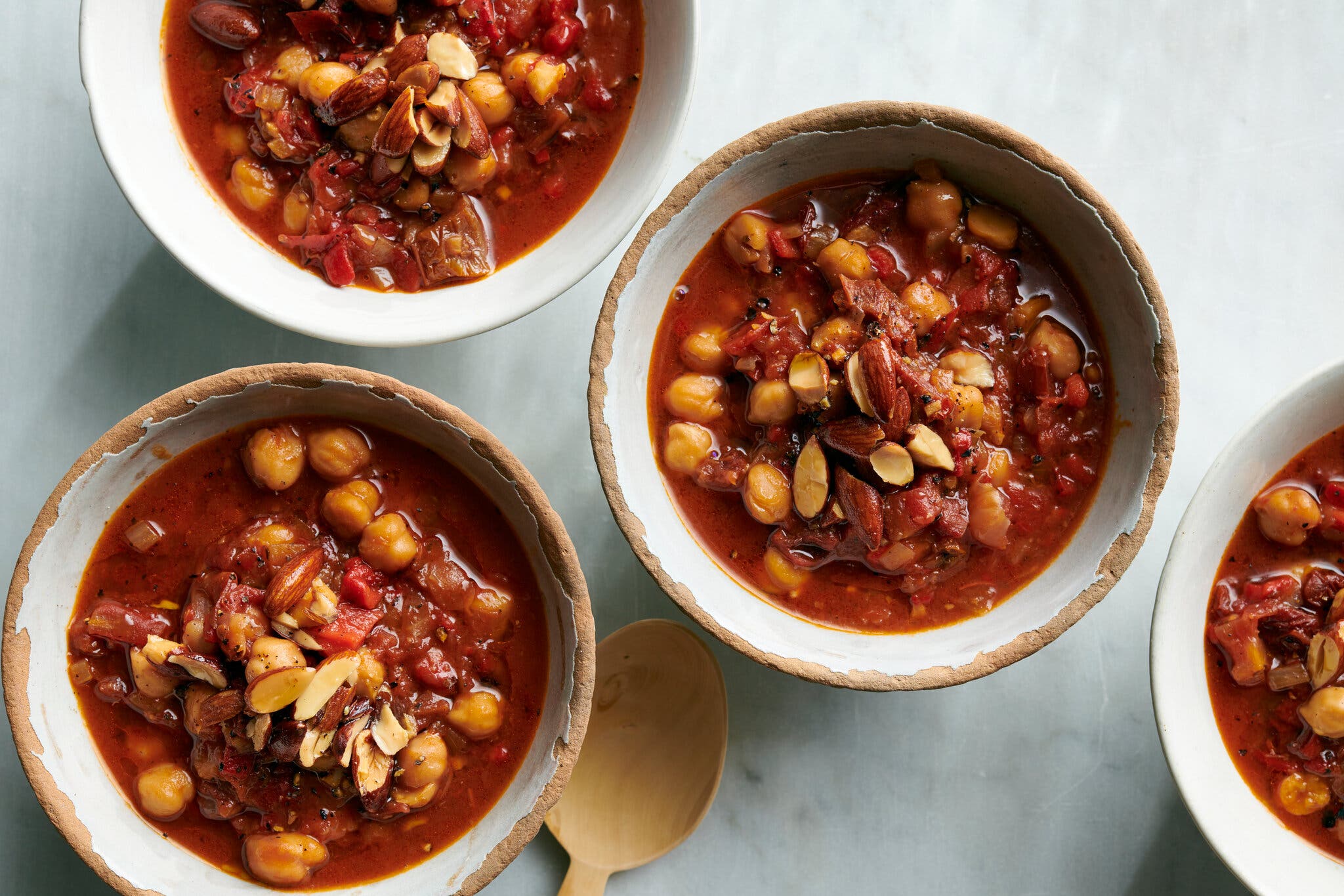 An overhead image of four rough-edged ceramic bowls of brick red tomato-chickpea stew scattered with smoked almonds.