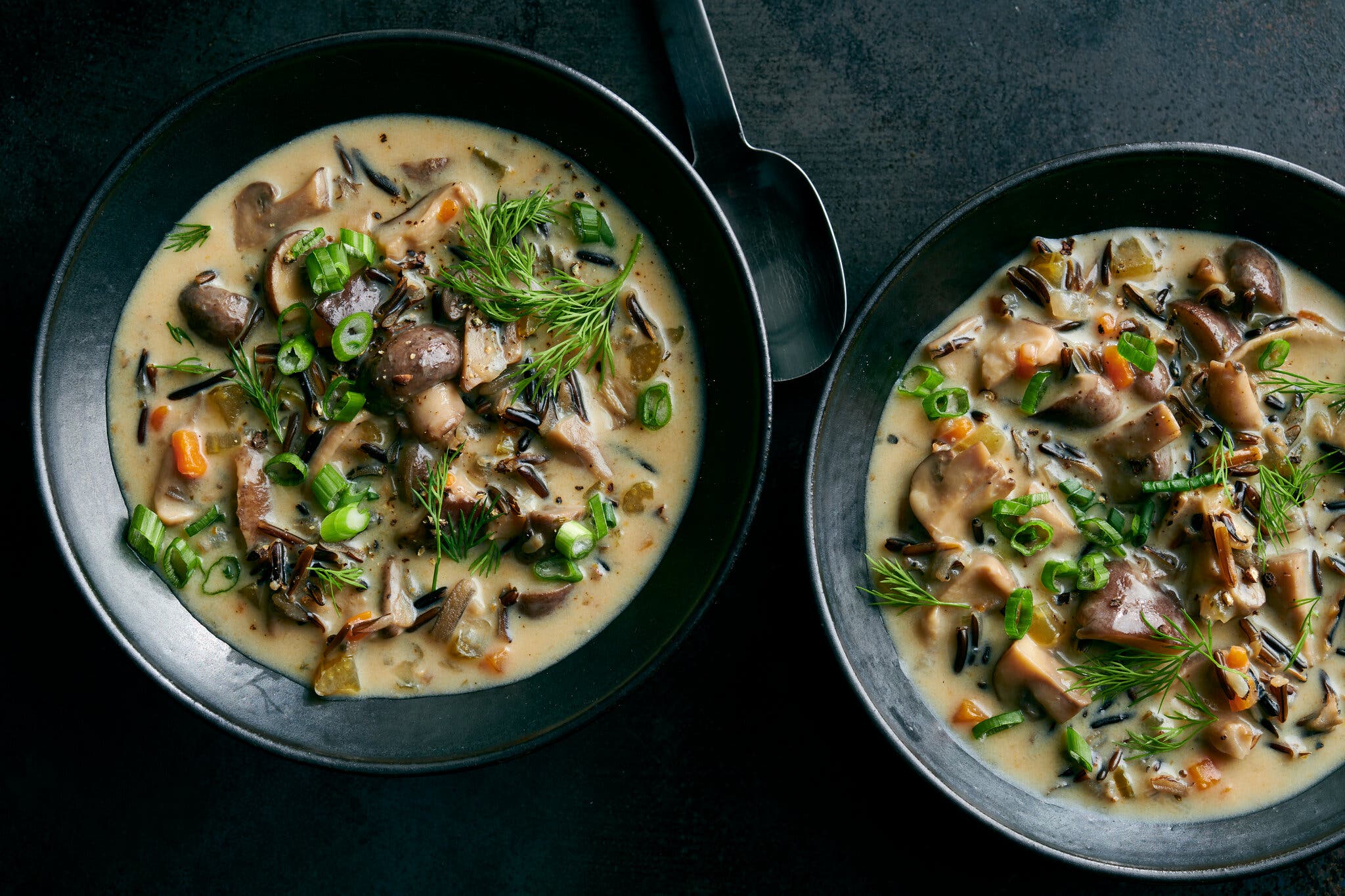 Two black bowls, sitting next to each other, are filled with a brothy soup punctuated with mushrooms. A smattering of green herbs sits on top.