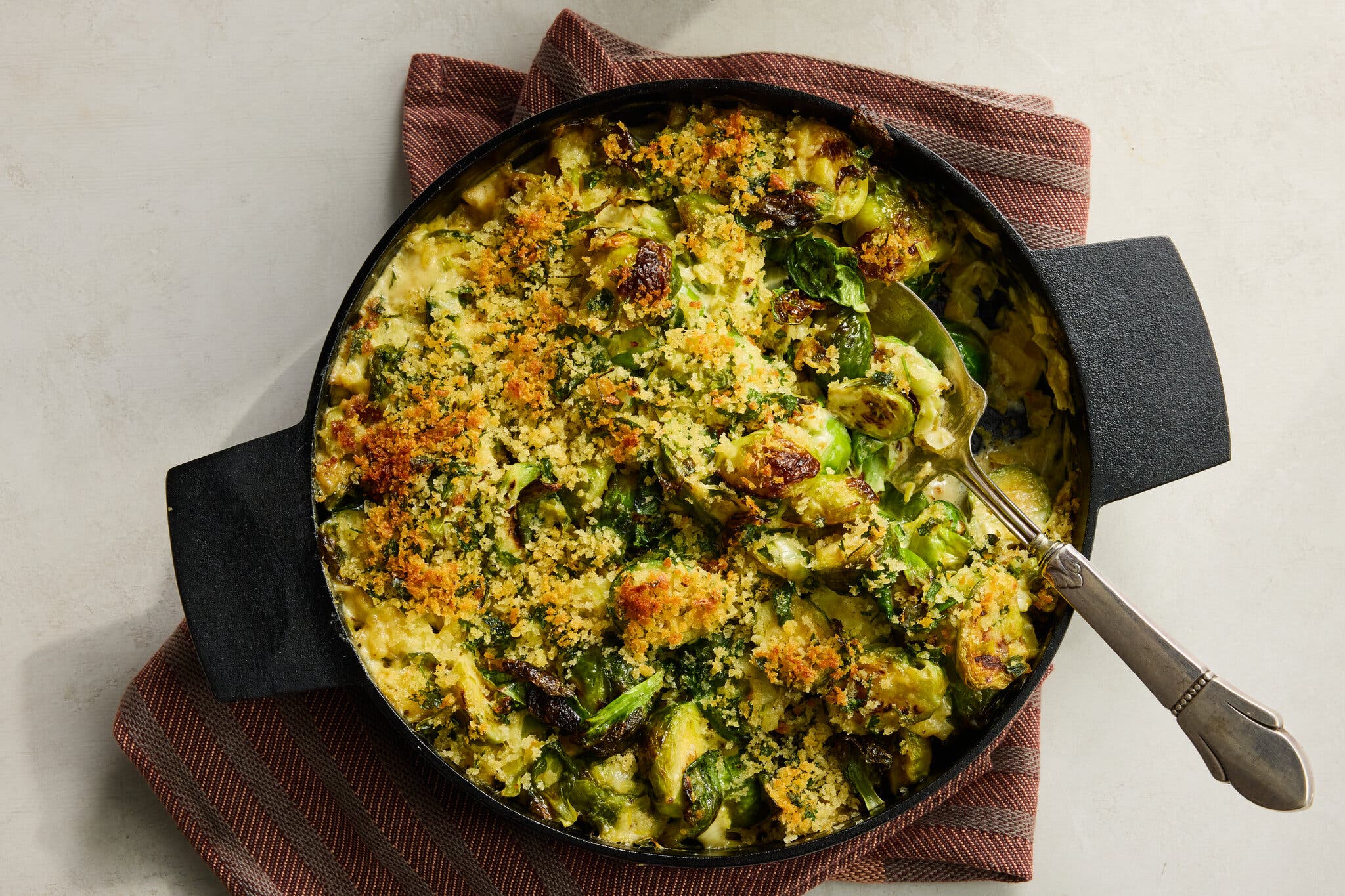 An overhead view of a brussels sprouts dish in a black cast-iron pan, topped with golden-brown breadcrumbs and served with a spoon.