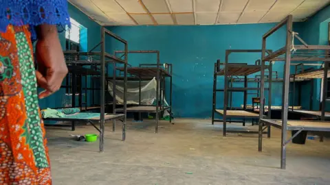 A woman's hand is seen to the left of the image as she looks on at empty black painted bunk beds in a dormitory at the school in Papiri - November 2025.