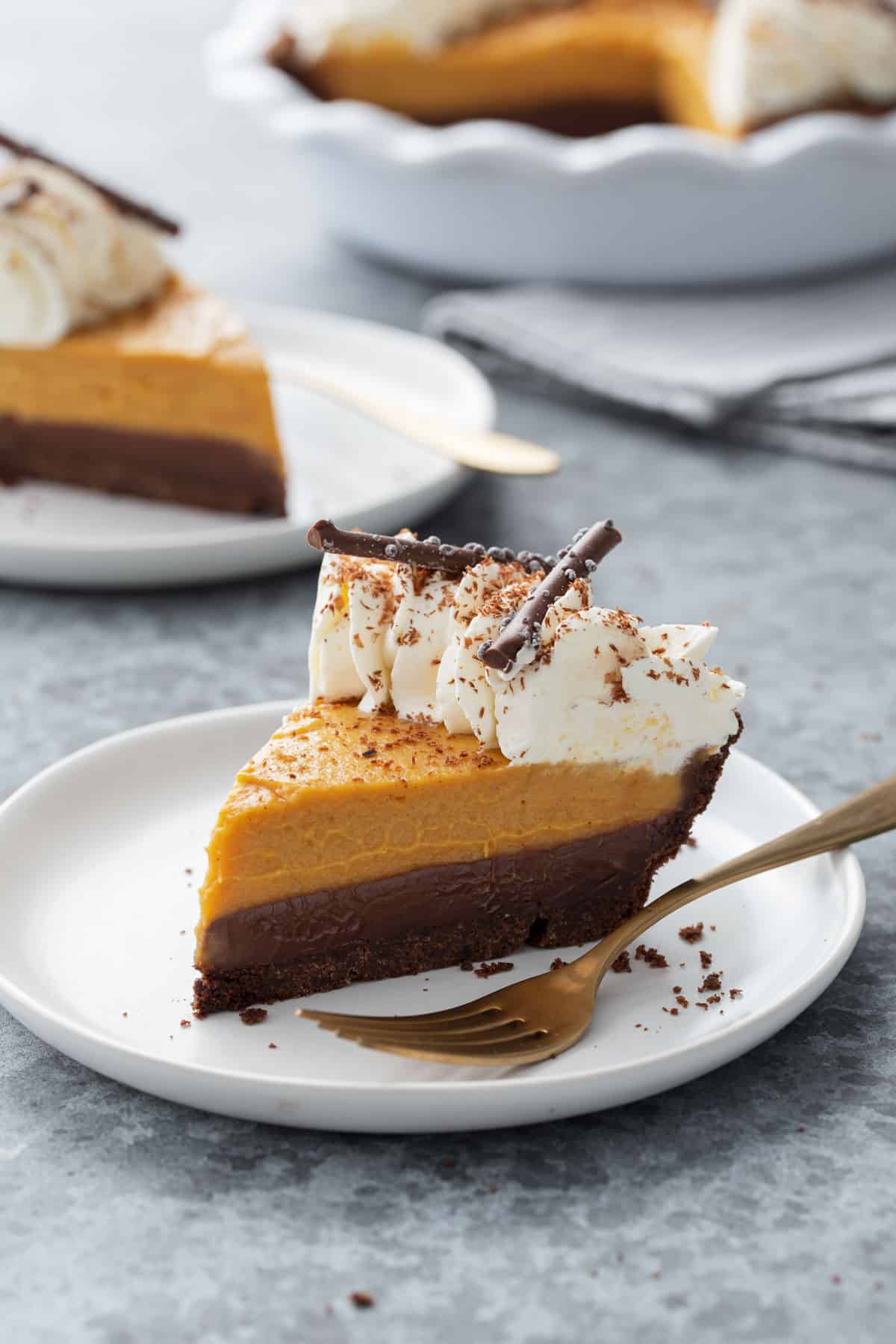 Closeup slice of Black Bottom Pumpkin Cream Pie on a white plate with gold fork, second slice and full pie dish in the background.
