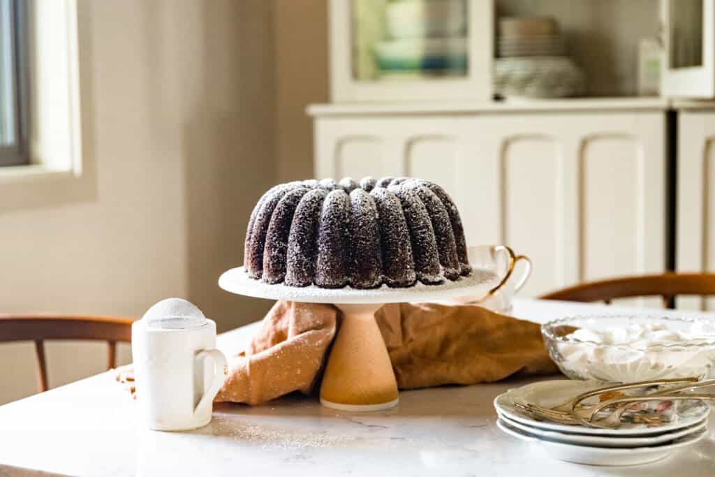 A Chocolate Bundt Cake on a Ceramic cake plate on top of a Marble countertop