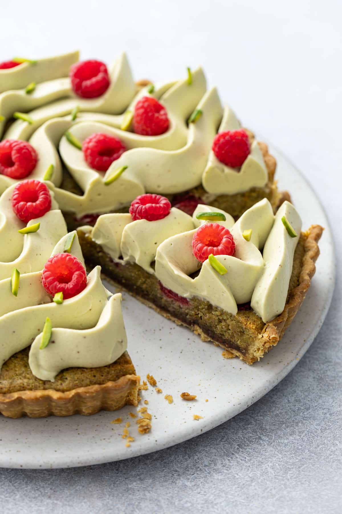 Raspberry & Pistachio Frangipane Tart with Whipped Pistachio Ganache on a flat cake plate, with one sliced piece to show the inside layers.