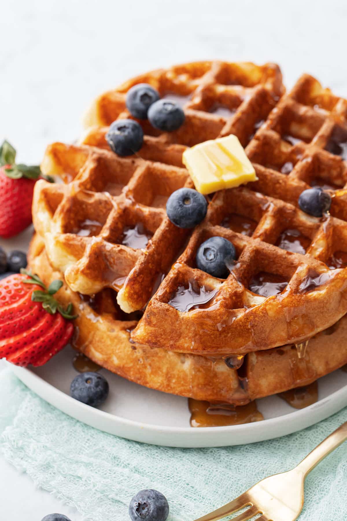 Plate with two golden brown sourdough Belgian waffles stacked and topped with berries, a pat of butter and a drizzle of syrup.